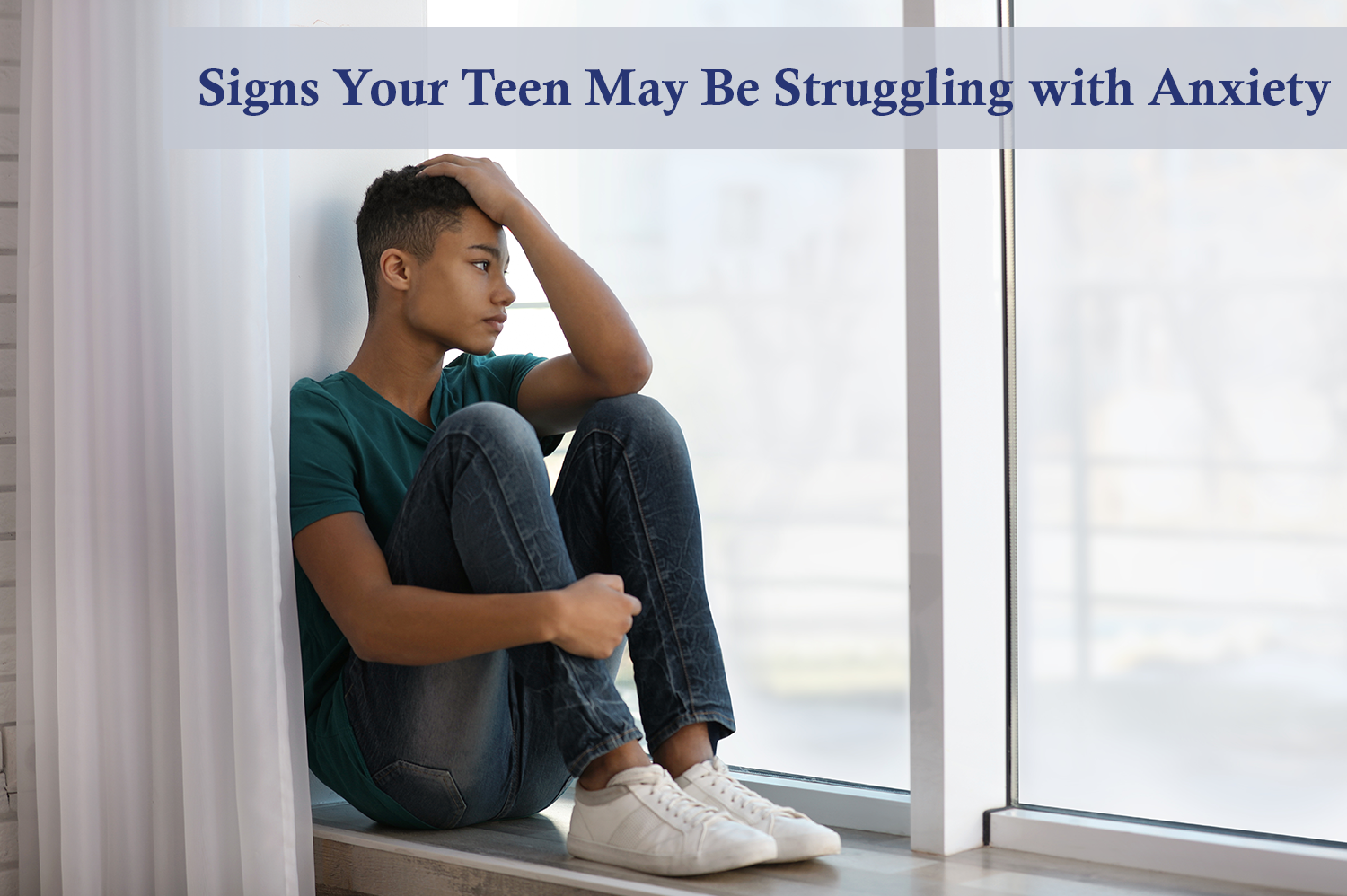Teen boy sitting in window ledge with hand in his hair, having anxiety.